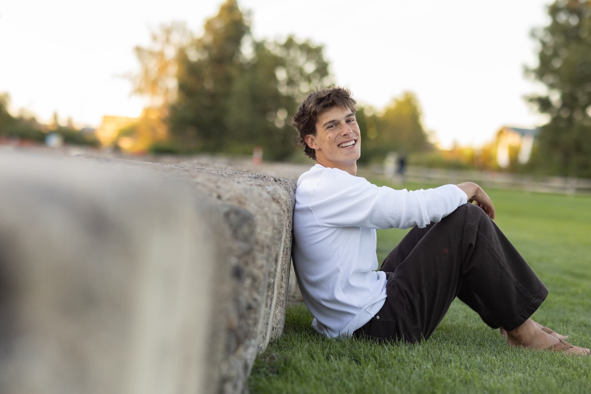 Senior guy laughing while leaning against a stone wall on green grass at sunset