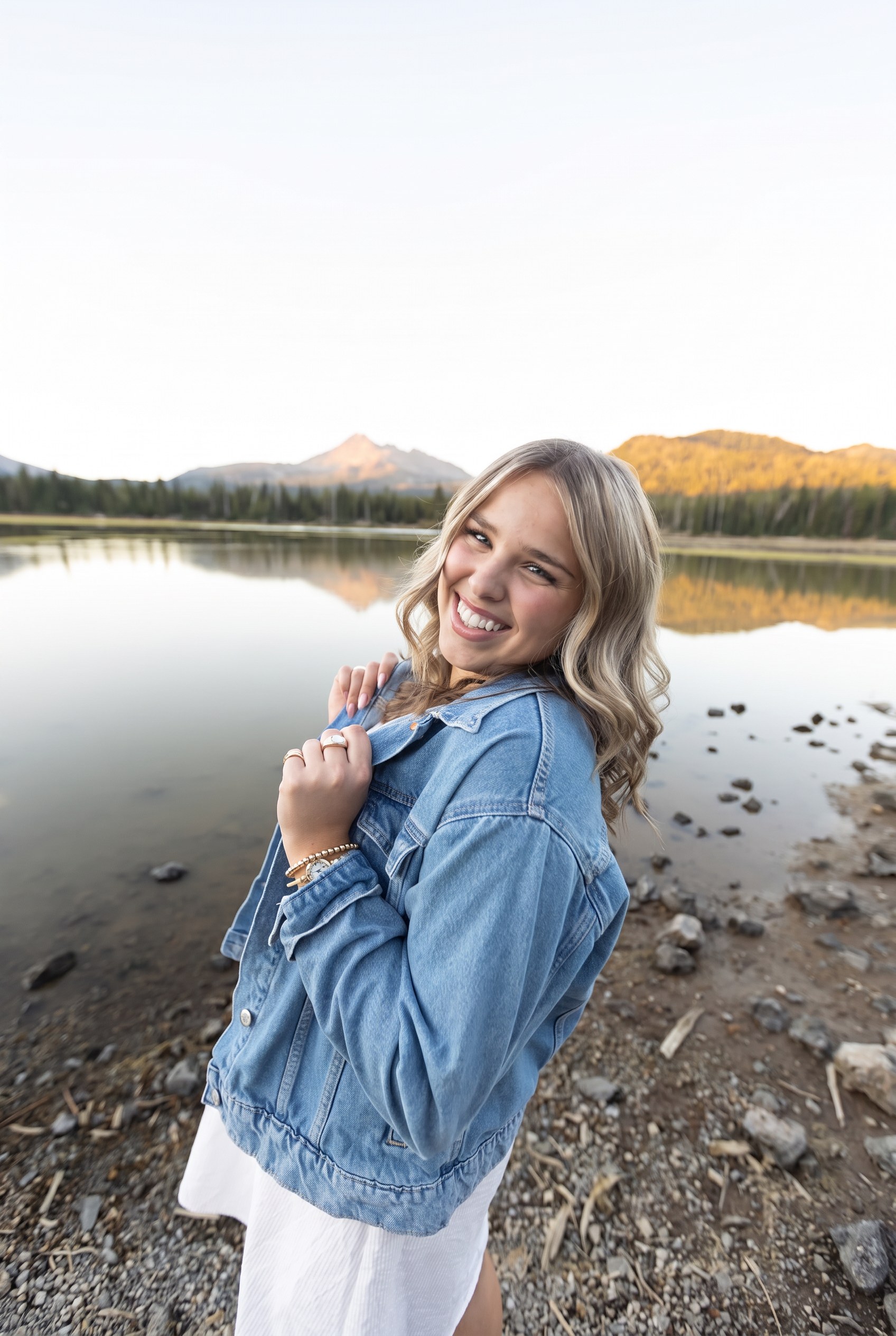 Senior girl in a denim jacket smiling by a mountain lake at sunset
