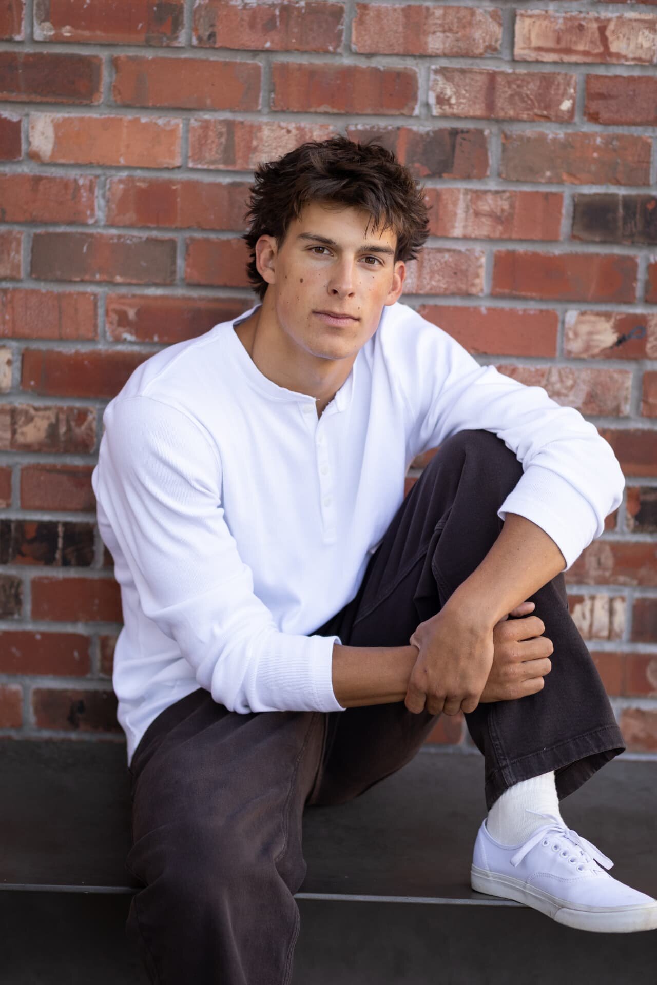 Senior guy in a white henley sitting against a red brick wall downtown