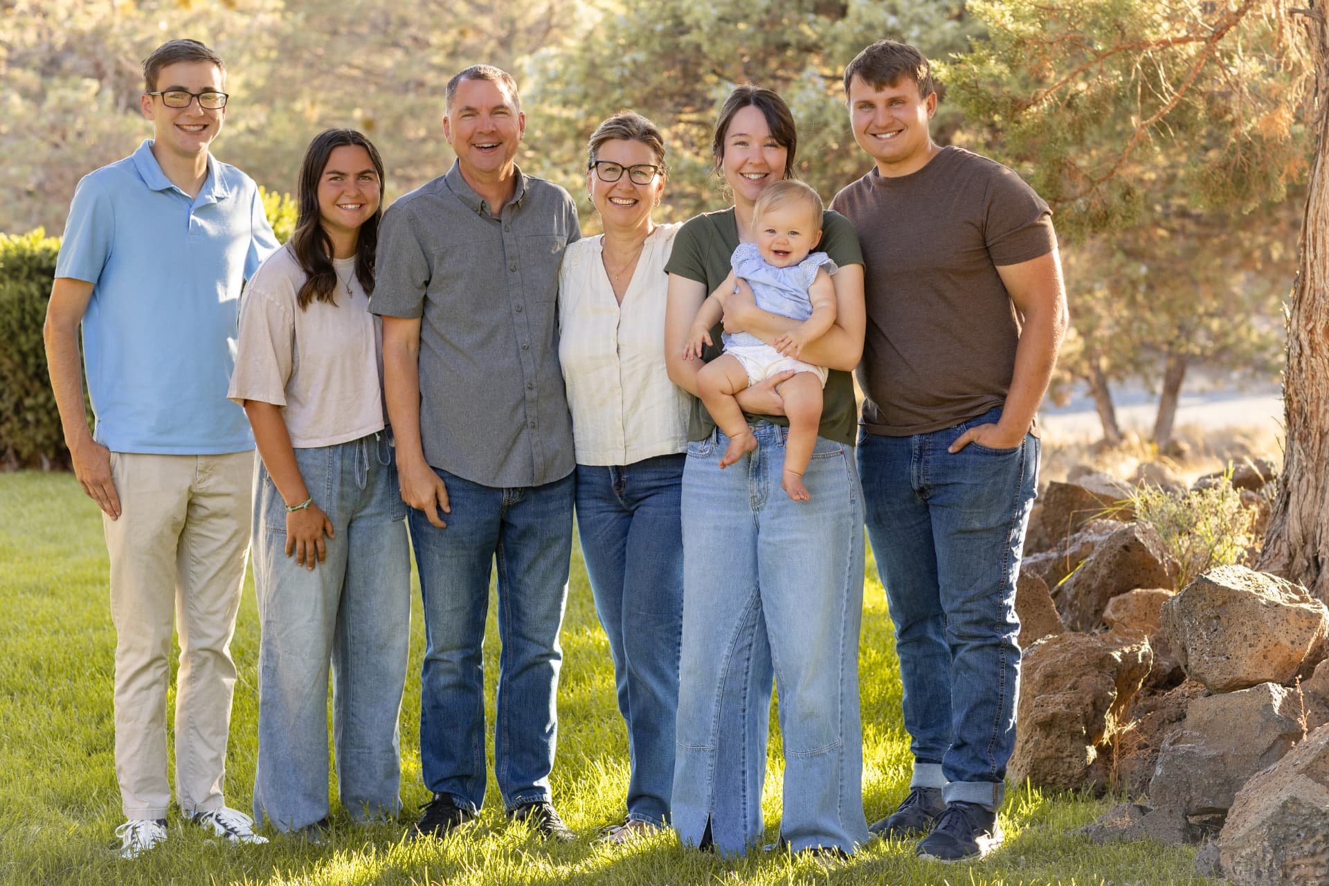 Extended family of seven laughing together on a sunlit lawn beneath junipers in Central Oregon