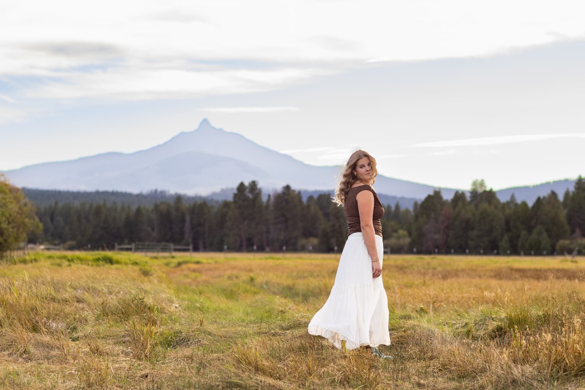 Senior in a white skirt standing in a meadow with a Cascade peak behind her