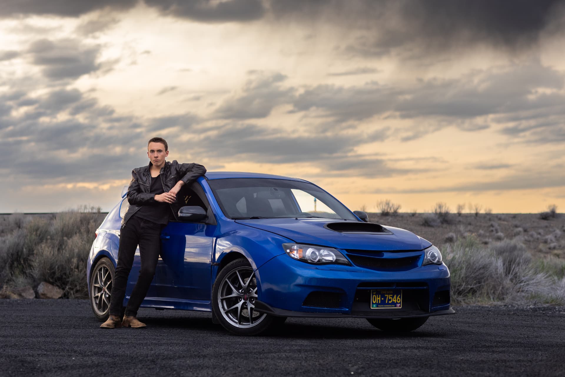 Senior guy leaning against his blue car at sunset in the high desert