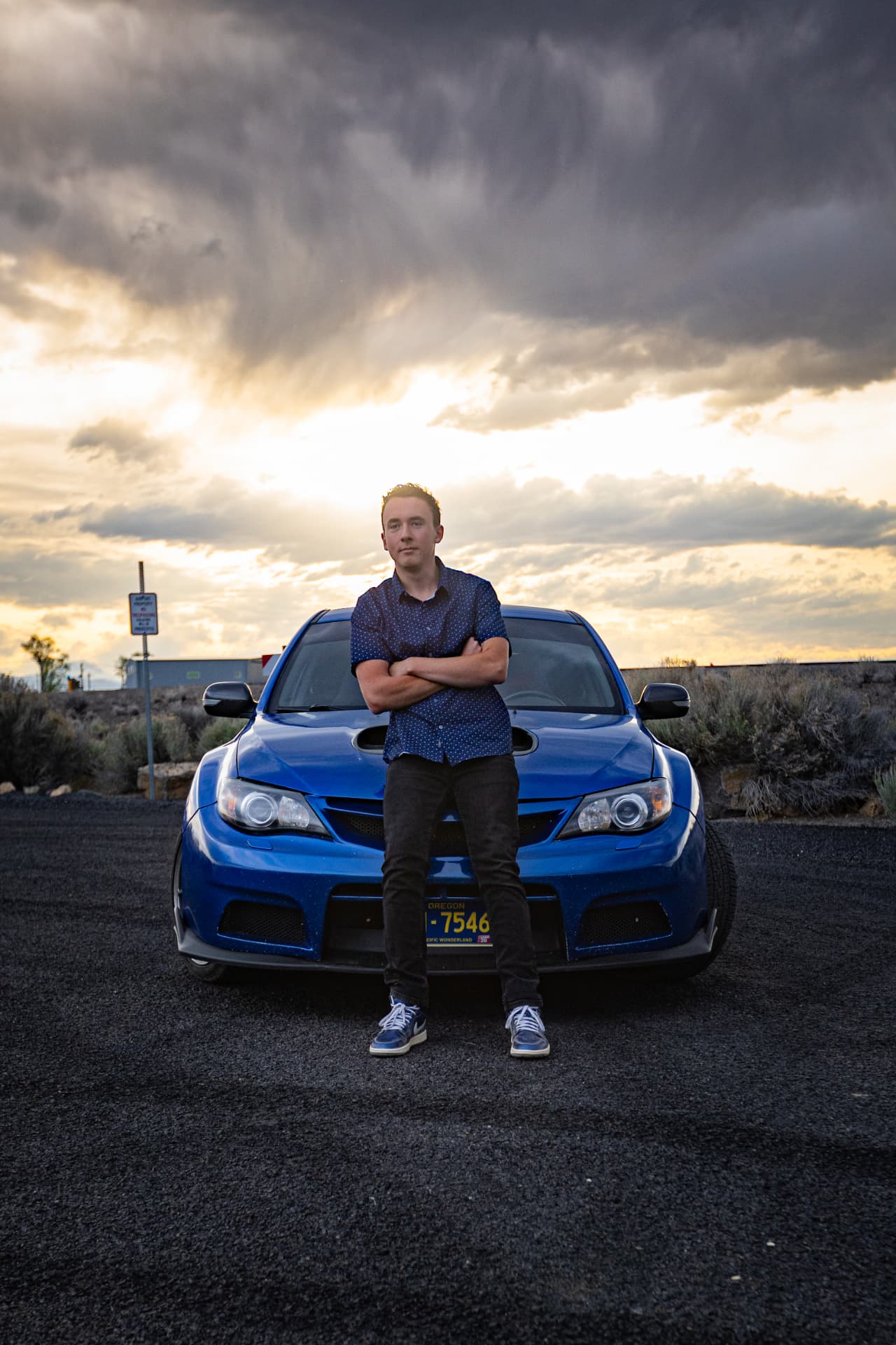 Senior guy leaning against the front of his blue car under dramatic clouds