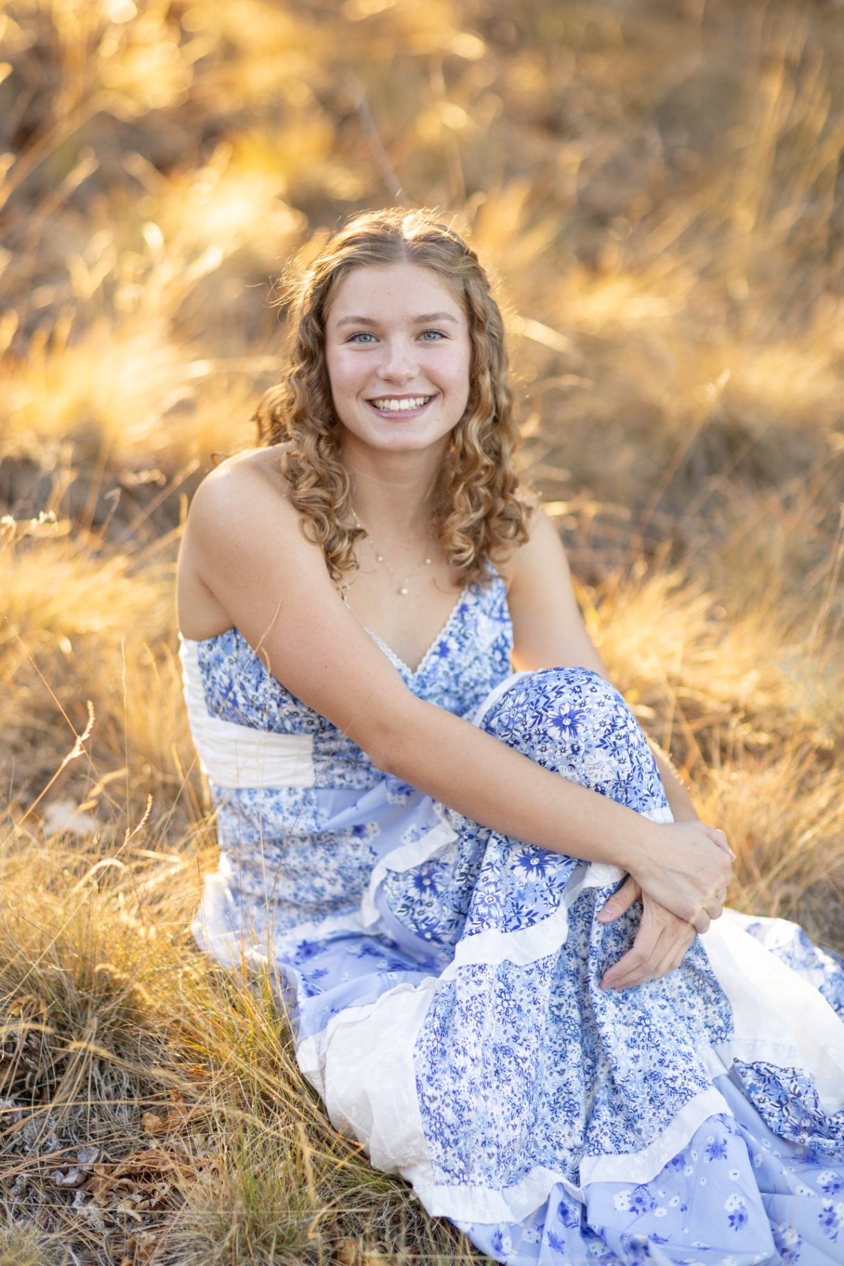 Senior girl in a blue floral dress smiling while seated in golden grass