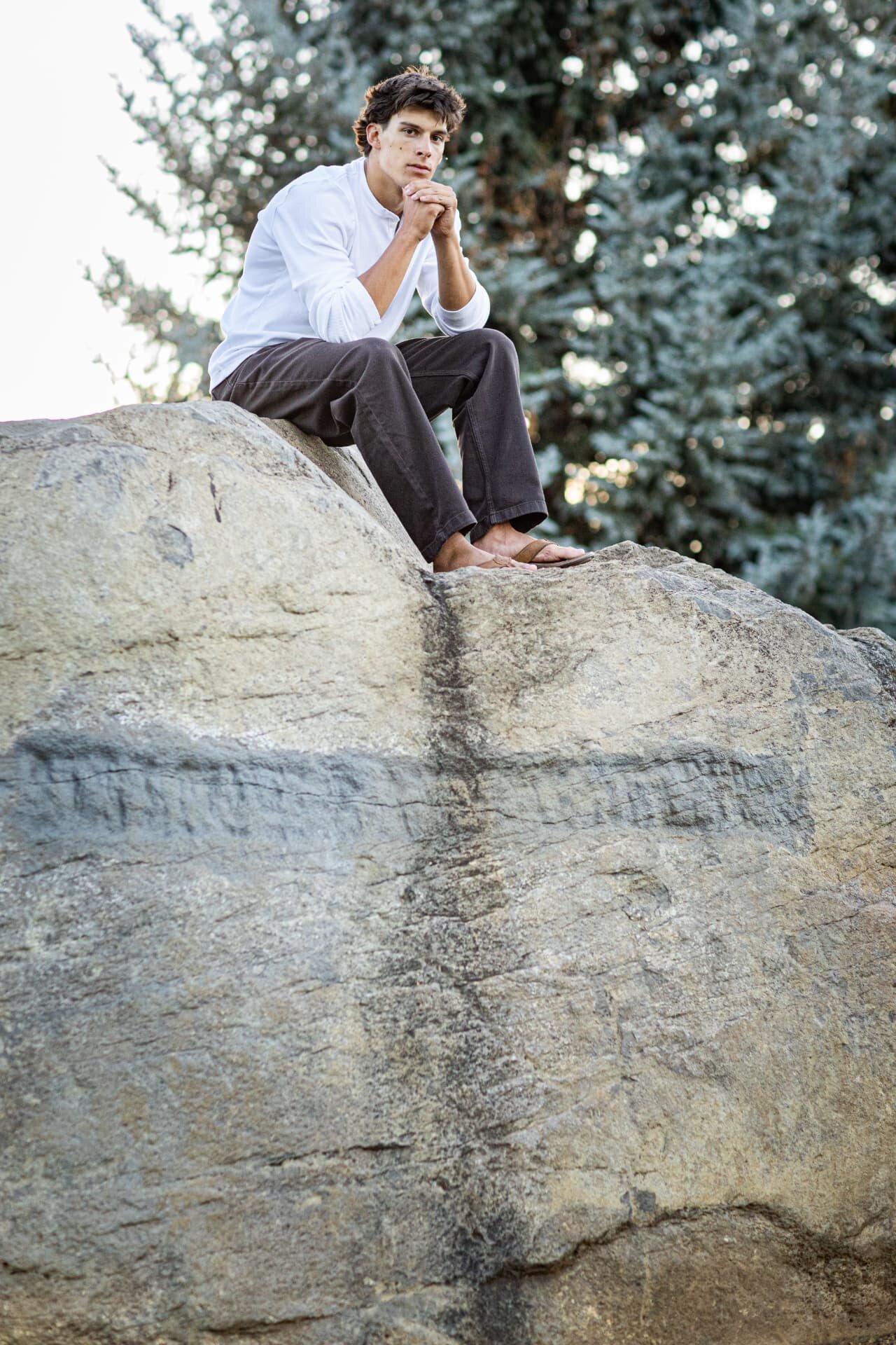 Senior guy sitting on a large granite boulder with evergreens behind him