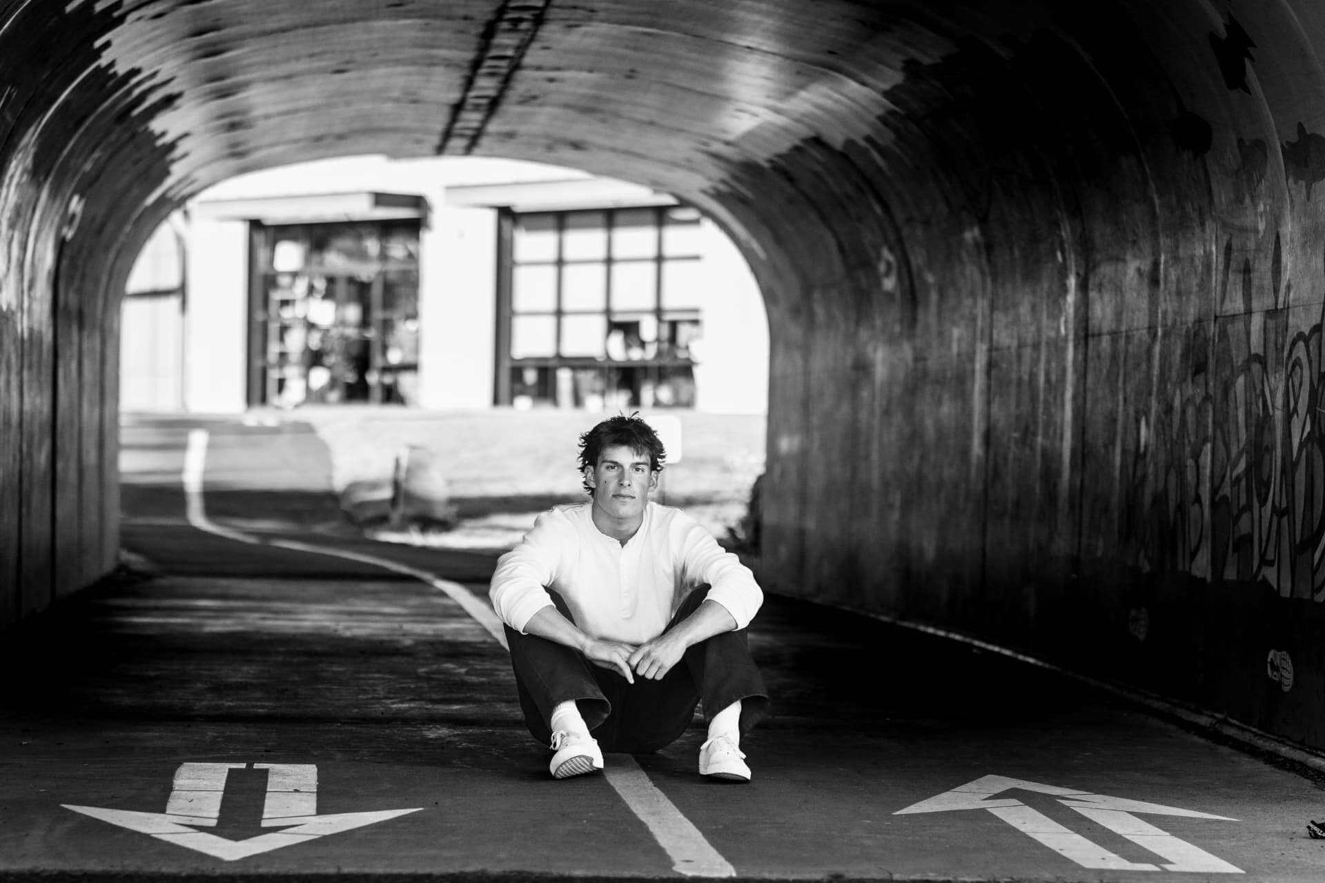 Black and white senior portrait of a guy sitting in a graffiti tunnel