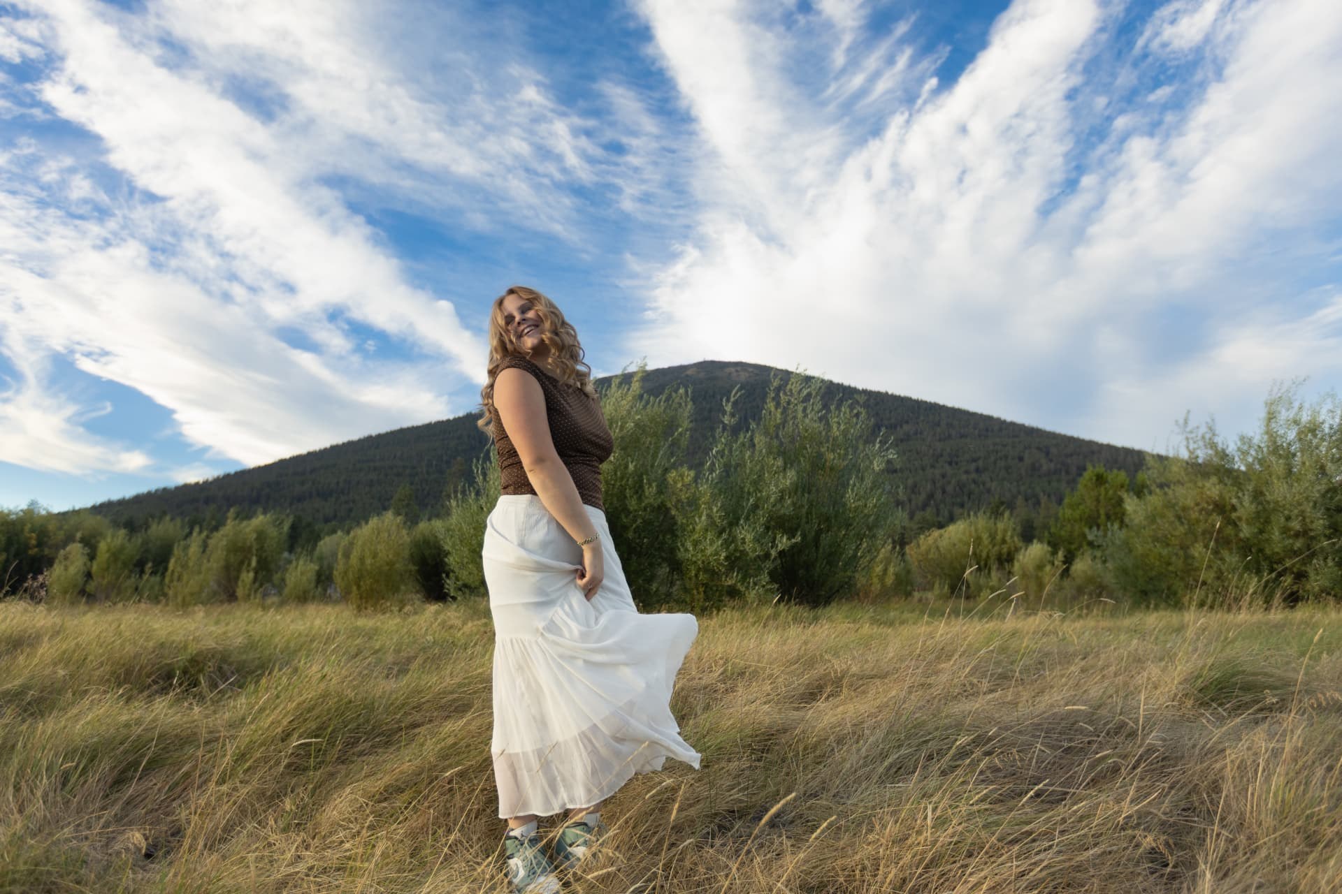 Senior girl spinning her skirt in a grass field below a forested butte near Sisters