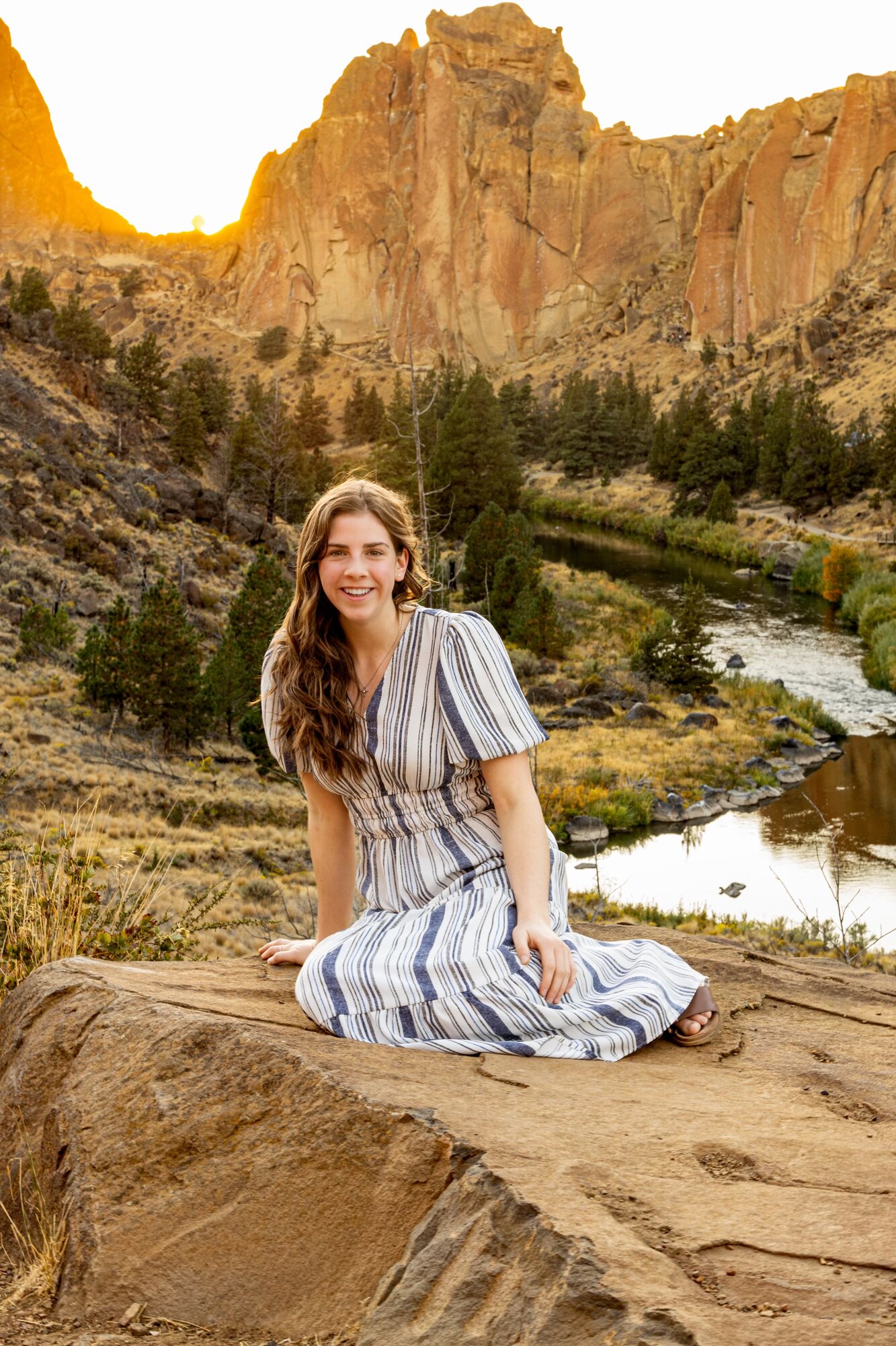 Senior girl in a striped dress seated on warm rock with Smith Rock behind her