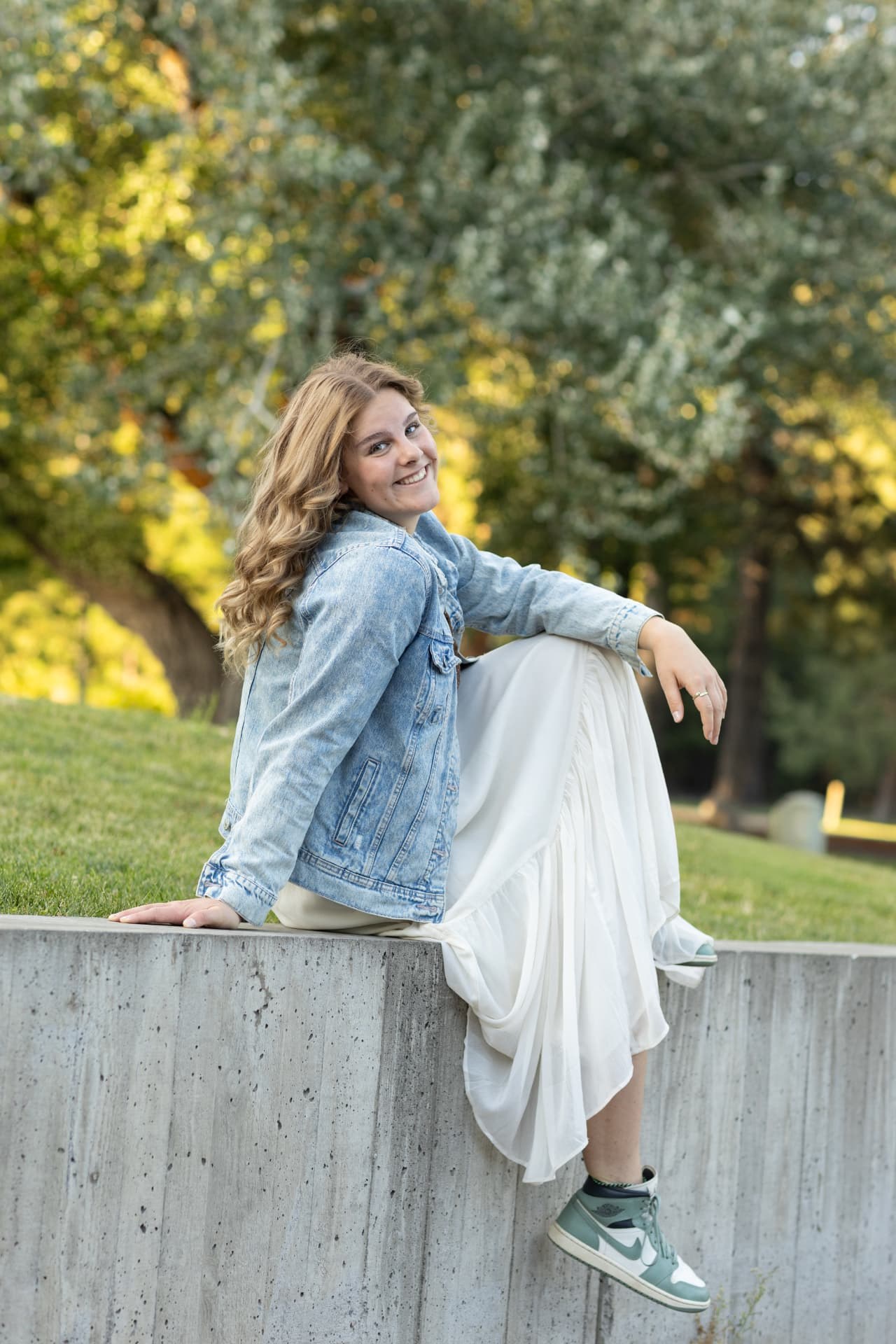 Senior girl in a denim jacket sitting on a concrete wall with sunlit trees behind her