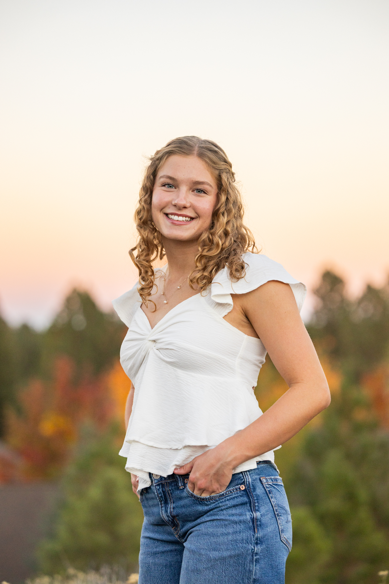 Senior girl smiling in a white top against a soft sunset sky