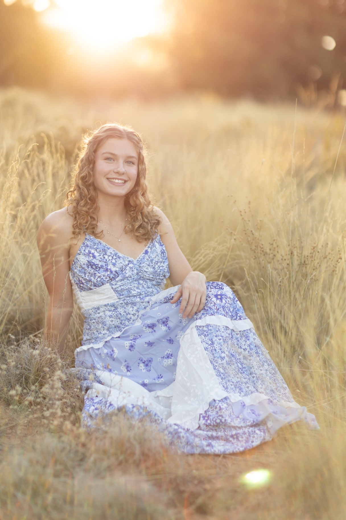 Senior girl in a blue floral dress seated in a sunlit meadow at sunset