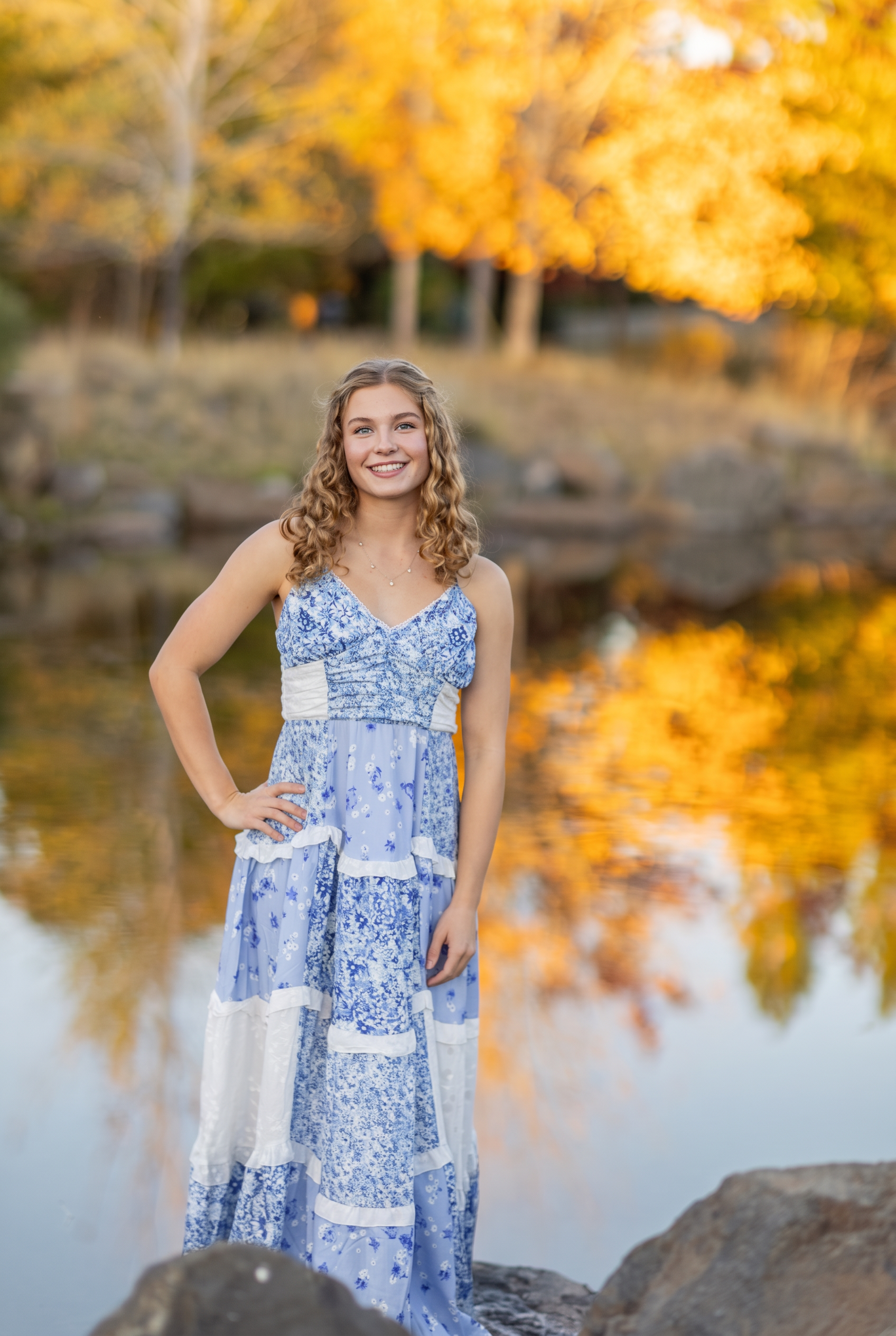 Senior girl in a blue floral dress beside a river reflecting golden autumn trees