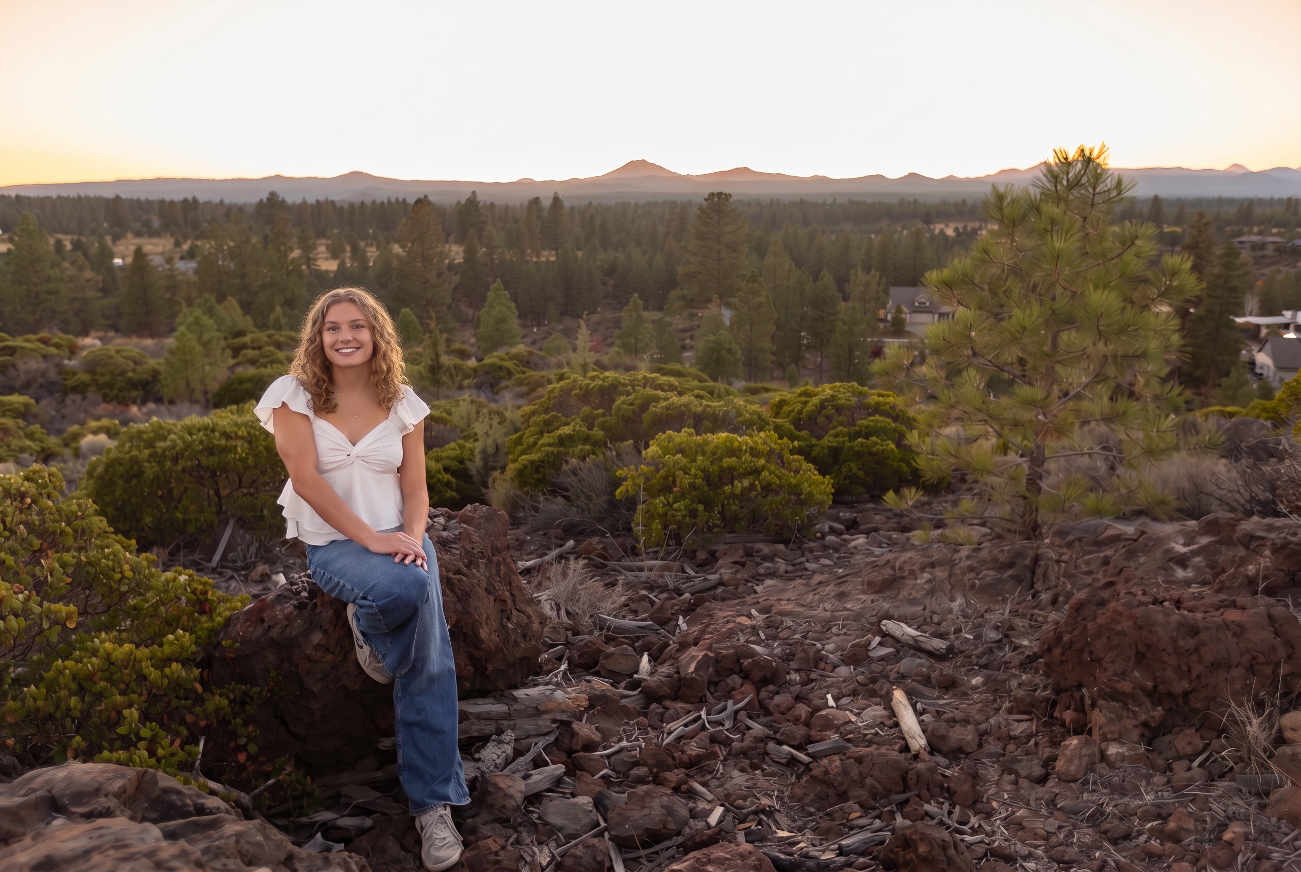 Senior girl in a white top seated on lava rock with the Cascade mountains on the horizon