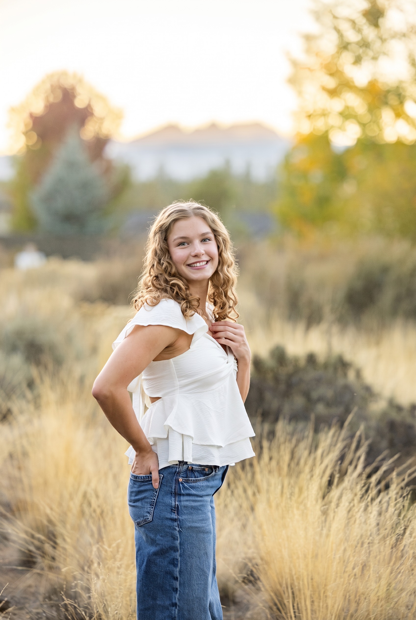 Senior girl in a white top and jeans standing in sunlit meadow grass